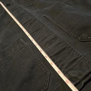 May include: Close-up of a black jacket with multiple pockets and button closures. A white measuring tape is laid across the jacket, showing measurements up to 23 inches.