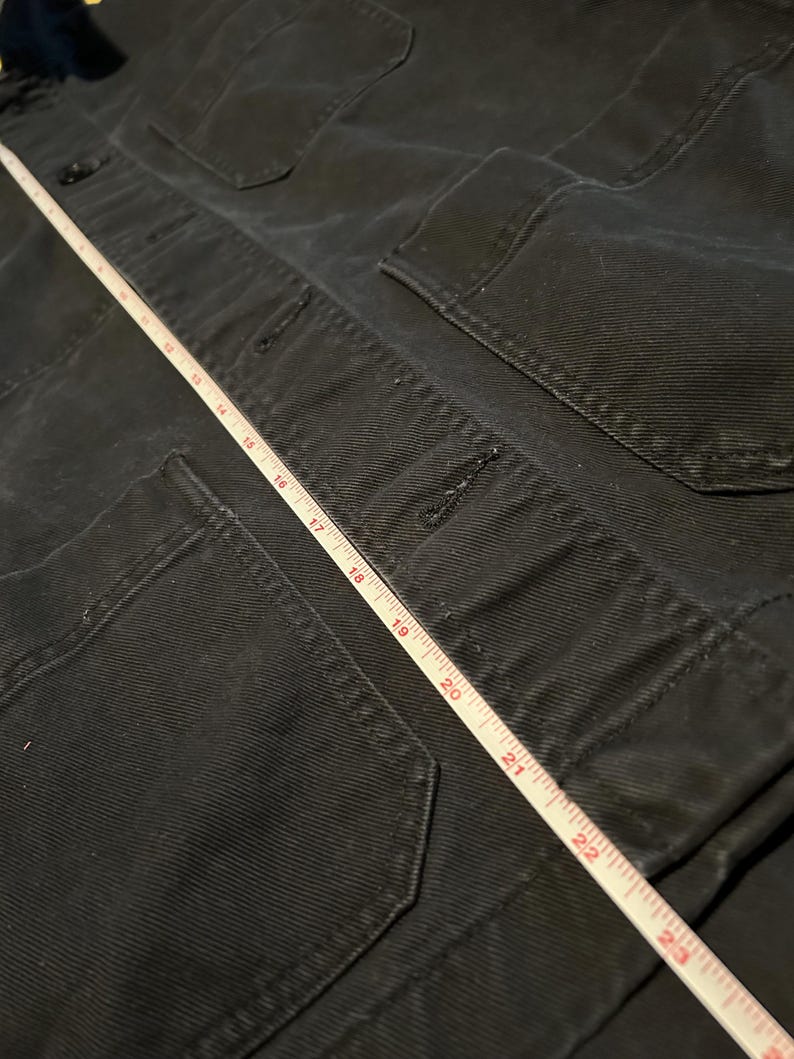 May include: Close-up of a black jacket with multiple pockets and button closures. A white measuring tape is laid across the jacket, showing measurements up to 23 inches.
