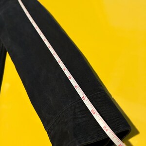 May include: A black jacket sleeve with a white measuring tape laid across it. The tape shows measurements up to 26 inches. The jacket is made of a dark fabric, possibly denim or a similar material.