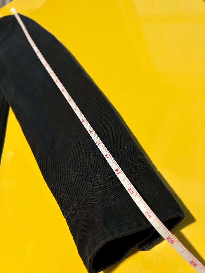 May include: A black jacket sleeve with a white measuring tape laid across it. The tape shows measurements up to 26 inches. The jacket is made of a dark fabric, possibly denim or a similar material.
