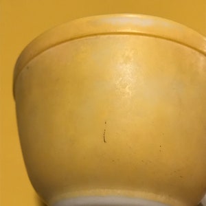 Könnte beinhalten: Eine hellgelbe Rührschüssel mit abgerundeter Form. Die Schüssel hat eine leicht strukturierte Oberfläche und einen weißen Rand. Die Schüssel besteht wahrscheinlich aus Glas oder Keramik und ist für die Zubereitung von Speisen bestimmt. Ein klassisches Küchenutensil.