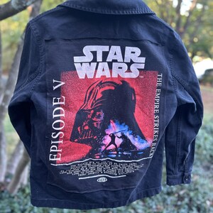 May include: Black denim jacket featuring a Star Wars Episode V graphic on the back. The graphic includes the Star Wars logo, Darth Vader, and the text "The Empire Strikes Back". The jacket has a button-up front and collar.