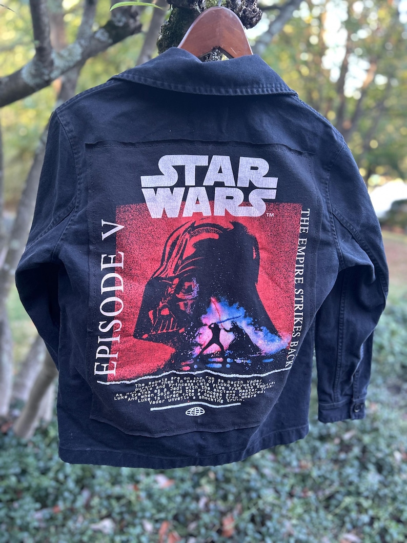 May include: Black denim jacket featuring a Star Wars Episode V graphic on the back. The graphic includes the Star Wars logo, Darth Vader, and the text "The Empire Strikes Back". The jacket has a button-up front and collar.