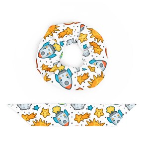 May include: A white scrunchie with a repeating pattern of cartoon rockets, stars, suns, and comets. The scrunchie is made of a soft, stretchy fabric.