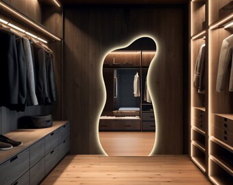 Op de afbeelding: Een moderne walk-in closet met houten panelen en warme verlichting. De kast heeft een grote, onregelmatig gevormde spiegel en planken met kleding die aan hangers hangen.