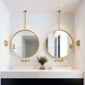 May include: A modern bathroom with two round, gold-framed mirrors suspended from the ceiling. The mirrors reflect the room's features, including white sinks, gold fixtures, and a black vanity. A small vase with greenery sits between the sinks.