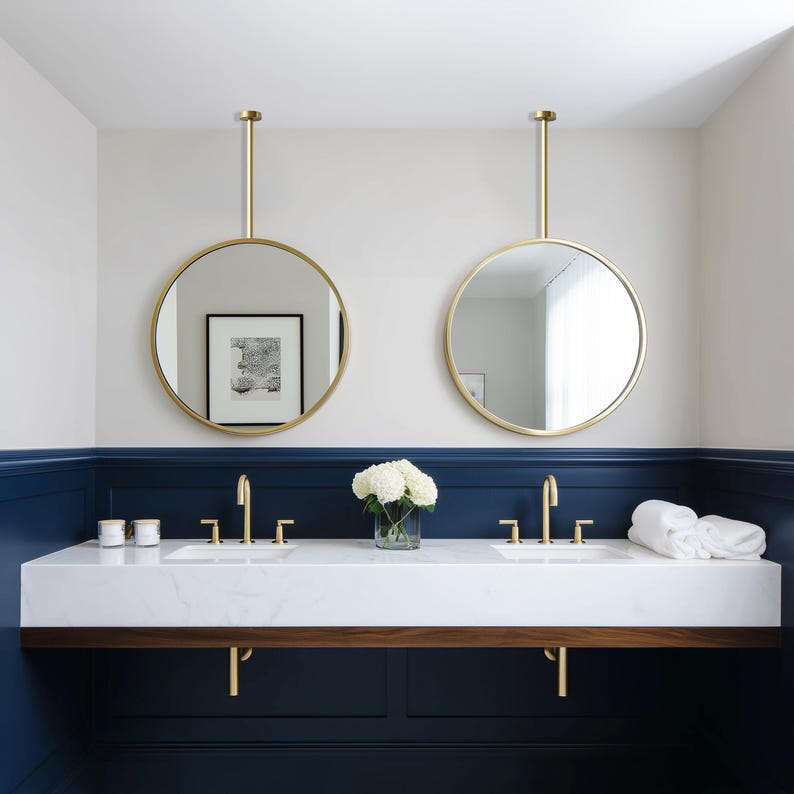 May include: A modern bathroom design featuring two round, gold-framed mirrors suspended from the ceiling. The mirrors hang above a white marble countertop with two sinks and gold faucets. The wall is painted navy blue and white.