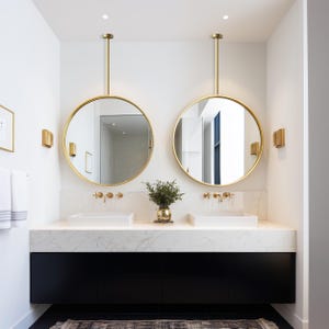 May include: A modern bathroom with two round, gold-framed mirrors suspended from the ceiling. The mirrors reflect the room's features, including white sinks, gold fixtures, and a black vanity. A small vase with greenery sits between the sinks.