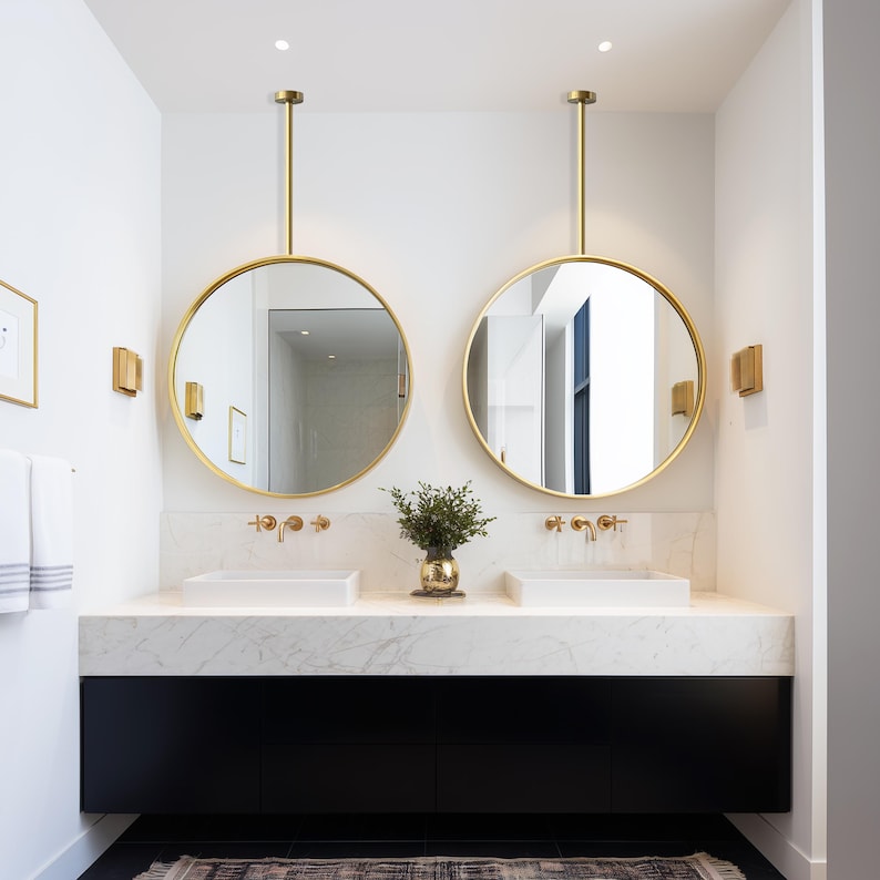 May include: A modern bathroom with two round, gold-framed mirrors suspended from the ceiling. The mirrors reflect the room's features, including white sinks, gold fixtures, and a black vanity. A small vase with greenery sits between the sinks.