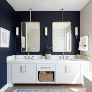 Op de afbeelding: Een moderne badkamer met een marineblauwe muur en witte kasten. Twee goudomrande spiegels hangen boven de dubbele wastafel. De wastafel heeft gouden armaturen en een rieten mand voor opslag.