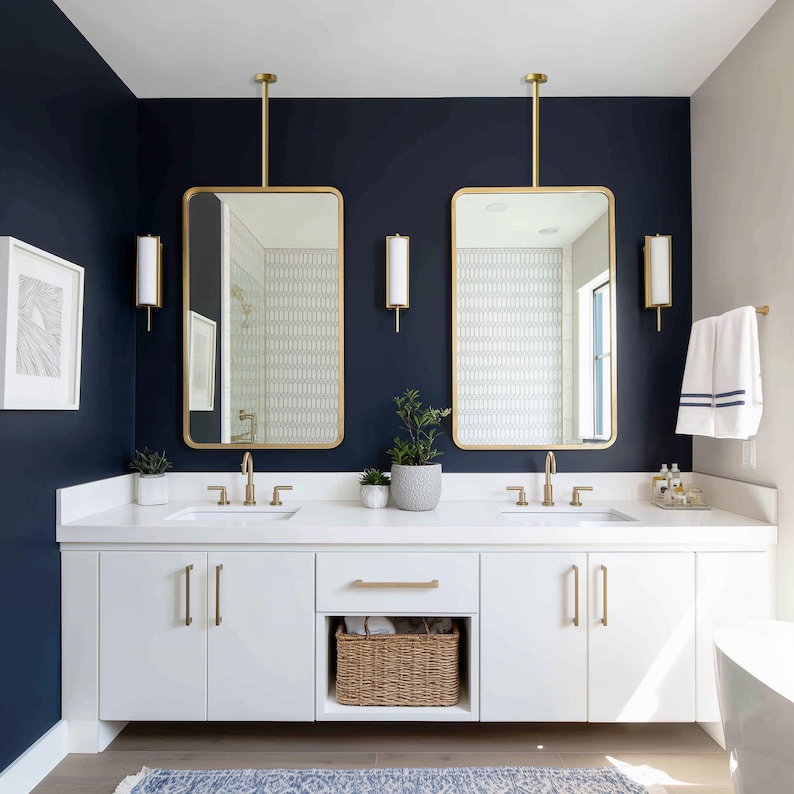 Op de afbeelding: Een moderne badkamer met een marineblauwe muur en witte kasten. Twee goudomrande spiegels hangen boven de dubbele wastafel. De wastafel heeft gouden armaturen en een rieten mand voor opslag.