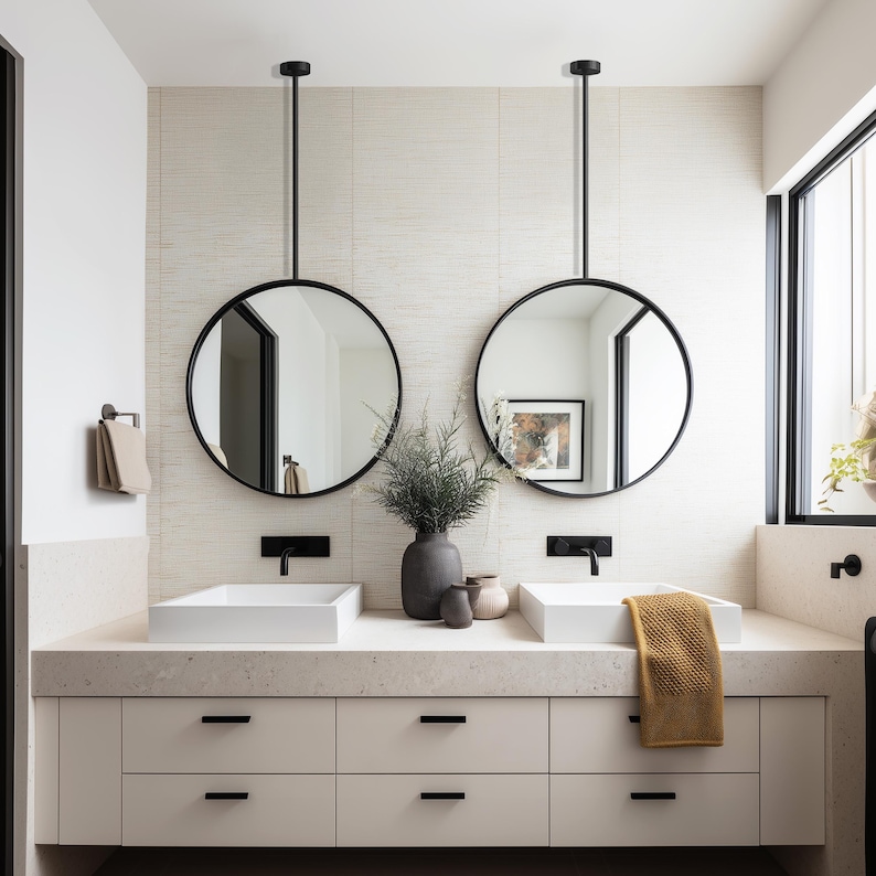 May include: A modern bathroom with two round, black-framed mirrors suspended from the ceiling. The mirrors reflect the room's interior, including white sinks, a beige countertop, and a white cabinet with black handles. A yellow towel hangs on the counter.