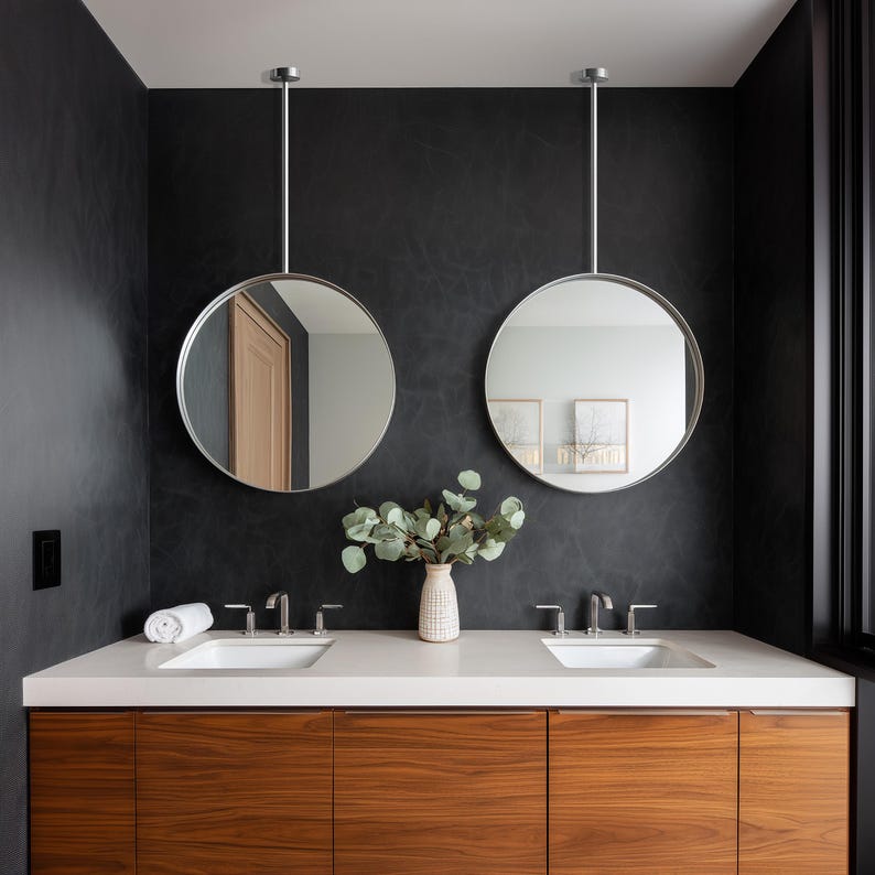 May include: A modern bathroom with a dark gray textured wall. Two round, silver-framed mirrors hang from the ceiling above a white countertop with two sinks. A wooden cabinet with horizontal graining sits below the countertop. A vase of greenery adds a natural touch.