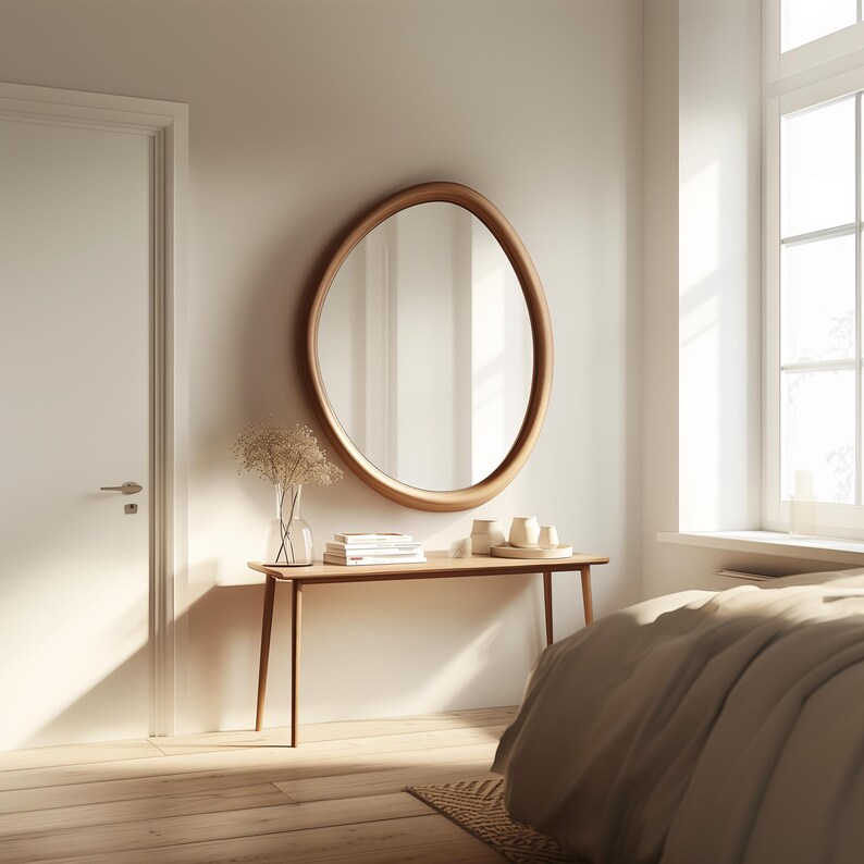 May include: A white bedroom with a wooden console table and a large oval mirror with a wooden frame. The table has a stack of books and decorative items on it. A bed with a beige duvet is in the background.