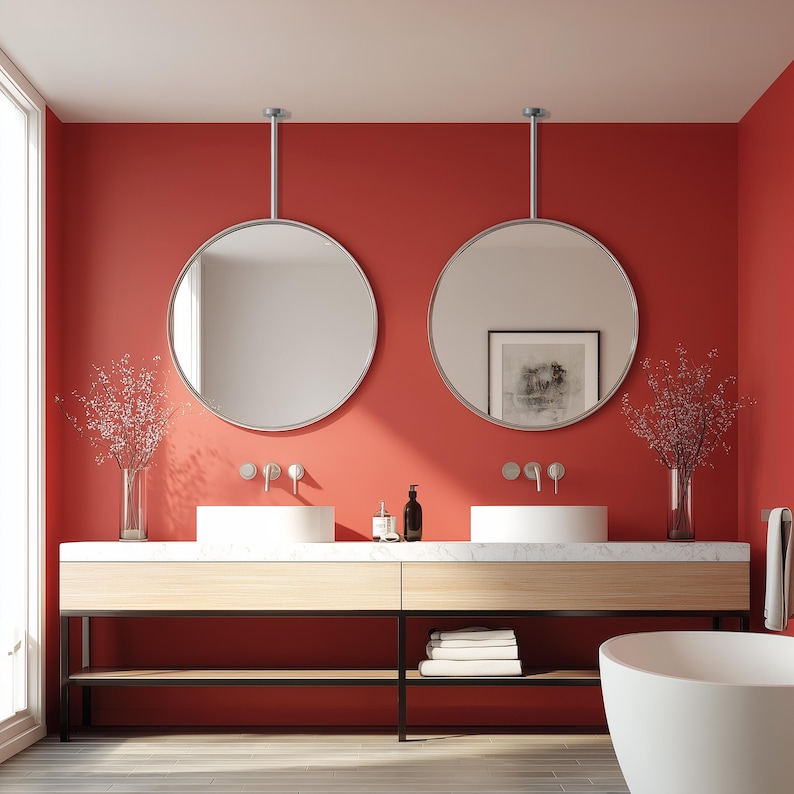 May include: A modern bathroom with a red wall, featuring two round mirrors suspended from the ceiling. The vanity has a light wood finish with a marble countertop and two white sinks. Decorative vases with floral arrangements and a white bathtub complete the scene.