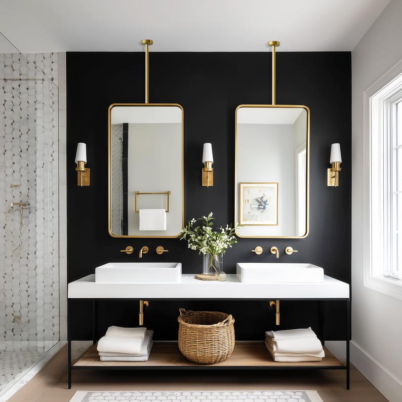 Op de afbeelding: Een moderne badkamer met een zwarte accentmuur, met twee rechthoekige spiegels met gouden lijst en witte wastafels. Gouden wandkandelaars en armaturen vullen het ontwerp aan. Een gevlochten mand en opgevouwen handdoeken voegen textuur toe. Een douche met een betegelde muur is zichtbaar.