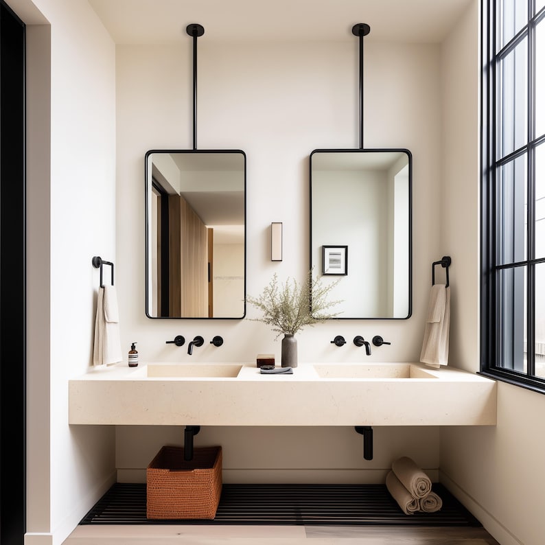 Op de afbeelding: Een moderne badkamer met twee rechthoekige spiegels die aan het plafond hangen. De spiegels hebben zwarte frames en bevinden zich boven een dubbele wastafel. De wastafel is gemaakt van lichtgekleurde steen, met zwarte kranen en een gevlochten mand.