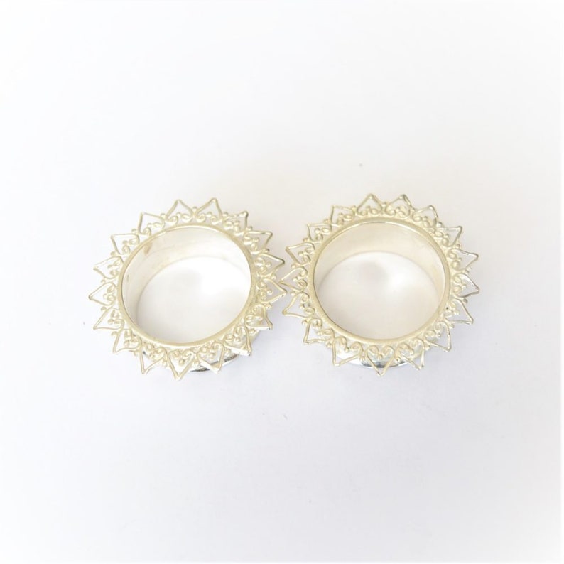 Puede incluir: Un par de dilatadores de oreja de plata con un dise&ntilde;o delicado e intrincado. Los dilatadores tienen una forma circular con un borde decorativo.