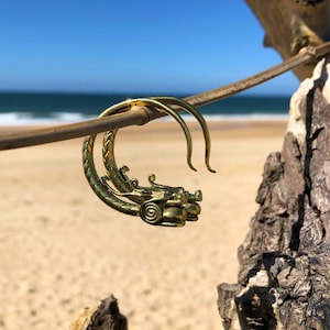 Può includere: Un paio di orecchini a cerchio dorati con un design a forma di drago. Gli orecchini sono appesi a un ramo su uno sfondo di spiaggia.