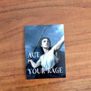 Könnte beinhalten: Ein rechteckiger Aufkleber mit einem klassischen Gemälde einer Person in einem weißen Gewand, die einen Speer vor einem dunklen, stürmischen Himmel hält. Der Text ACT YOUR RAGE ist in weißen Großbuchstaben im unteren Teil abgedruckt.