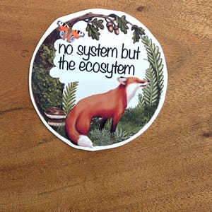 Könnte beinhalten: Ein runder Aufkleber mit einem Rotfuchs in einer Waldumgebung mit grünen Farnen, Pilzen, einem Eichenzweig mit Eicheln und einem Schmetterling. Der Text "no system but the ecosytem" ist in schwarzen Buchstaben auf weißem Grund gedruckt.