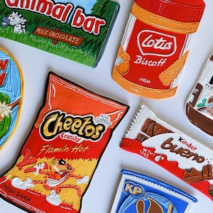 May include: A collection of colorful, hand-painted replicas of popular snack food packaging. The replicas include Cheeto's Flamin' Hot, Kinder Bueno, Nutella, Lotus Biscoff spread, Mandarin Crush, Animal Bar, and a generic brand of Cheez-Its.