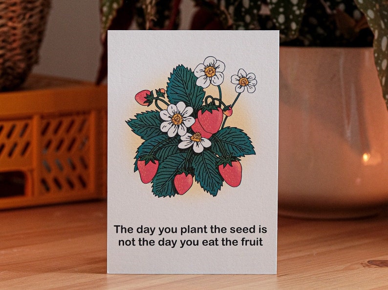 May include: A greeting card with a strawberry illustration, including red berries, green leaves, and white flowers. The card reads: "The day you plant the seed is not the day you eat the fruit."