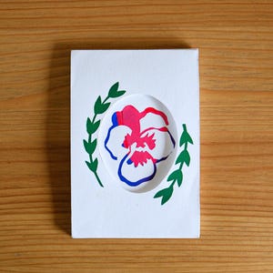 Könnte beinhalten: Eine weiße Karte mit einem ausgeschnittenen Blumendesign. Die Blume ist rosa, rot und blau, umgeben von grünen Blättern. Die Karte liegt auf einer Holzoberfläche.