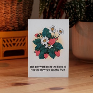 May include: A greeting card with a strawberry illustration. The design includes red strawberries, white flowers, and green leaves. The card reads: "The day you plant the seed is not the day you eat the fruit."