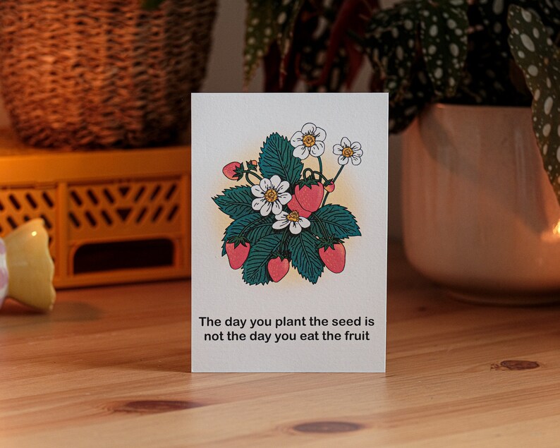 May include: A greeting card with a strawberry illustration. The design includes red strawberries, white flowers, and green leaves. The card reads: "The day you plant the seed is not the day you eat the fruit."