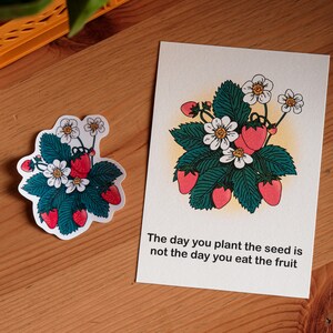 May include: A sticker and a card with a strawberry plant design. The illustration shows red strawberries, white flowers, and green leaves. The card reads: "The day you plant the seed is not the day you eat the fruit."