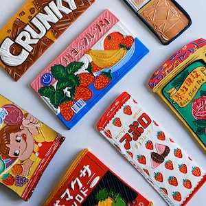 May include: Assortment of colorful chocolate bar wrappers with Japanese text. The wrappers feature illustrations of fruit, flowers, and cartoon characters. The text on the wrappers includes "Crunky", "耕生のフルーツ", "マクサ シロドー", "meg", "アポロ", "チョコレート", "茶紅瑰玫", and "Rose Black Tea".