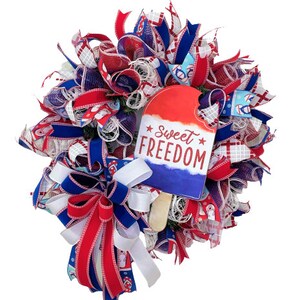 May include: A red, white and blue Fourth of July wreath with a popsicle-shaped sign that reads "Sweet Freedom". The wreath is made of mesh and ribbon and features a bow in the bottom left corner.
