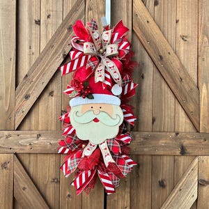 May include: A Christmas wreath featuring a central wooden Santa Claus face with a white beard and red hat. The wreath is decorated with red and white striped ribbons, plaid fabric, and red berry accents, all arranged in a vertical teardrop shape.