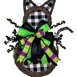 May include: A black and white checked cat-shaped wreath with a green and black bow. The wreath is made of twigs and has a black and white checked fabric covering the cat's body. The bow is made of green and black ribbon with purple and orange stripes.