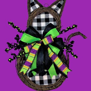 May include: A black and white checked cat-shaped wreath with a green and purple bow. The wreath is made of twigs and has black and white polka dot accents.