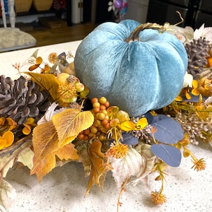 May include: A teal velvet pumpkin centerpiece with fall foliage, pine cones, and berries. The centerpiece is perfect for a Thanksgiving or autumn table decoration.