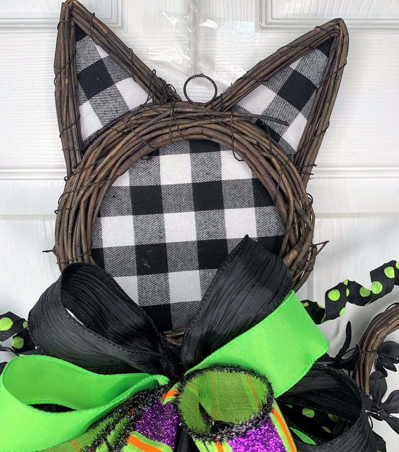 May include: Black and white buffalo check fabric wreath with black and green ribbon bow. The wreath is shaped like a cat's head with wire ears.