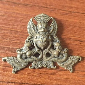 May include: A brass-coloured metal pendant featuring a central winged figure with a bird-like head, flanked by two serpent-like creatures with coiled tails. The design includes intricate decorative scrollwork at the base and top edges.