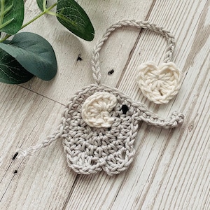 May include: A crocheted elephant ornament with a grey body and white ears. The elephant has a small black eye and is attached to a white crocheted heart.