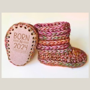 Puede incluir: Un par de botitas de bebé de crochet rosa, verde y naranja con una etiqueta de cuero que dice "Born in 2024 Limited Edition".