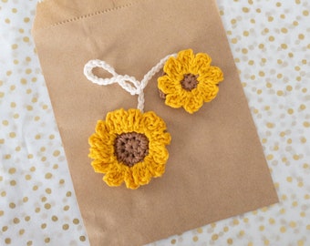 May include: Two crocheted sunflowers, one larger than the other, with brown centers and yellow petals, are tied together with white string and placed on a brown paper bag.