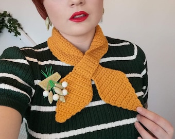 May include: A woman with short red hair is wearing a green and white striped sweater with a yellow crocheted scarf tied around her neck. She is wearing a white and gold brooch with a green stem and white berries.
