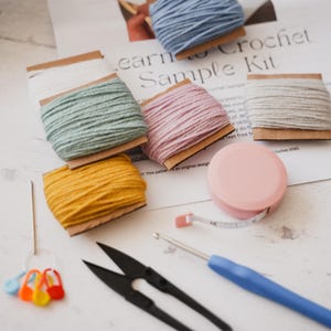 A flat lay of a complete learn to crochet kit, including yarn, hook, scissors, tape measure, pompom maker and PDF print out of the pattern for a hat with pompom.