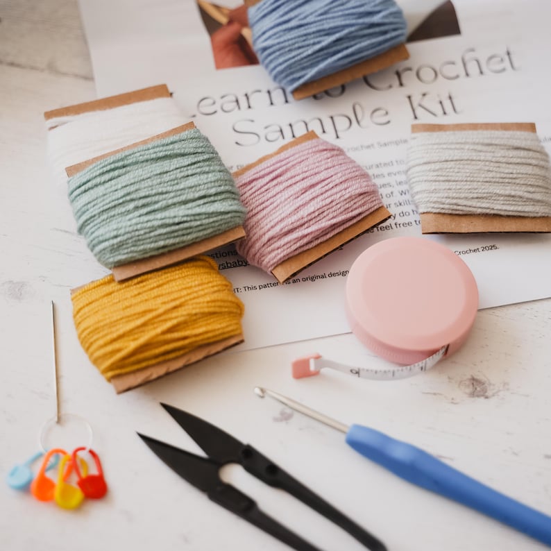 A flat lay of a complete learn to crochet kit, including yarn, hook, scissors, tape measure, pompom maker and PDF print out of the pattern for a hat with pompom.