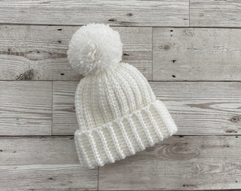 May include: White knitted beanie with a fluffy pom pom on top. The beanie is made of a soft, textured yarn and has a ribbed design.
