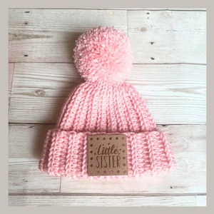 May include: A light pink knitted beanie with a large pom-pom on top. The hat features a brown square patch with the words "Little Sister" in a decorative font. The beanie is made of a chunky knit material.
