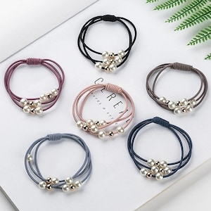 May include: Six hair ties with pearl and gold accents. The hair ties are in various colors, including black, purple, brown, pink, blue, and gray.