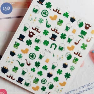 May include: A sheet of white nail stickers with a variety of St. Patrick's Day themed designs. The designs include shamrocks, beer mugs, leprechaun hats, pipes, horseshoes, and boots. The stickers are perfect for adding a festive touch to your nails.