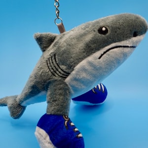 May include: A plush gray shark keychain with blue sneakers. The shark has a white belly, black gills, and a sad expression. The keychain has a silver ring and a red strap.